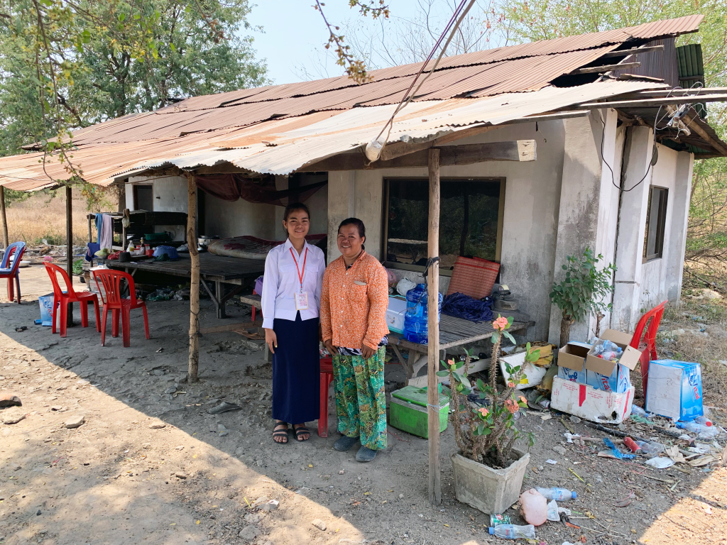 Sothea and her mom stand in front of a dilapidated house smiling with holes in the roof and debris everywhere